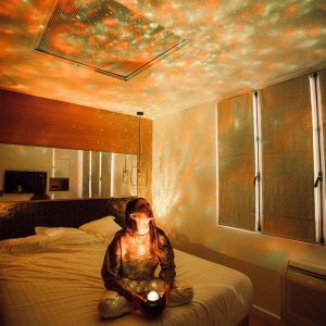 Vrouw die op een bed zit in een kamer, een sterrenbol gebruikt om lichtpatronen op de muren en het plafond te projecteren, waardoor een ontspannende en magische sfeer ontstaat.