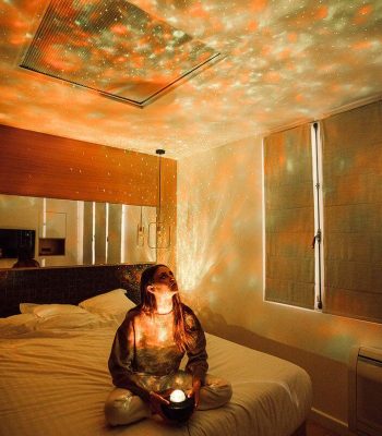 Vrouw die op een bed zit in een kamer, een sterrenbol gebruikt om lichtpatronen op de muren en het plafond te projecteren, waardoor een ontspannende en magische sfeer ontstaat.
