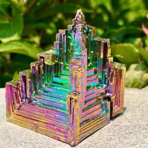 Decoratief stuk van iriserend metaal, geïnspireerd op een piramidevormige geometrische structuur, met kleurrijke reflecties, geplaatst op een rotsachtige ondergrond buiten.